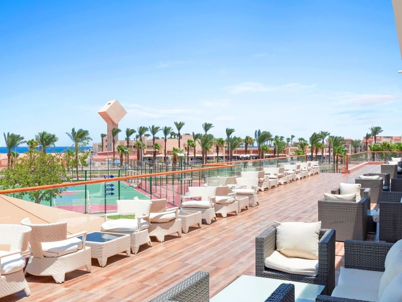 Outdoor lounge area with wicker chairs and tables, overlooking a tennis court and palm trees.