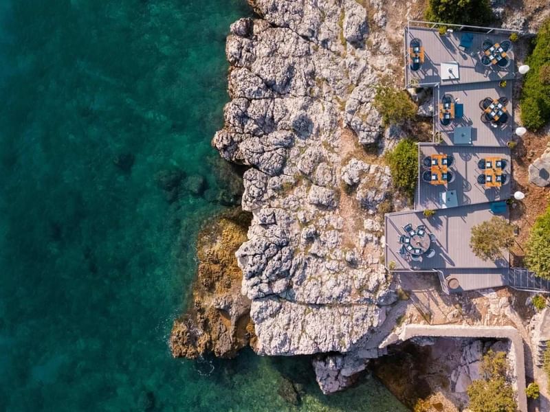 Luftaufnahme eines Restaurant-Terrasses am Meer mit blauem Wasser und felsigem Ufer.