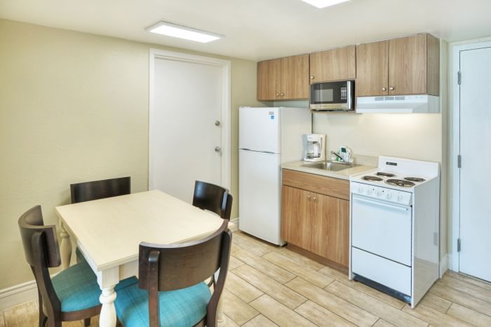 A compact kitchen with a dining area, featuring a small kitchenette with white appliances, a dining table with four chairs, and light wood flooring.