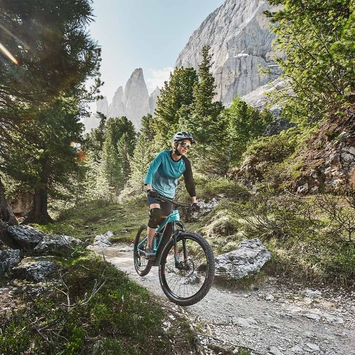 Ciclista in mountain bike su sentiero sterrato in bosco montano con rocce e sole tra gli alberi.