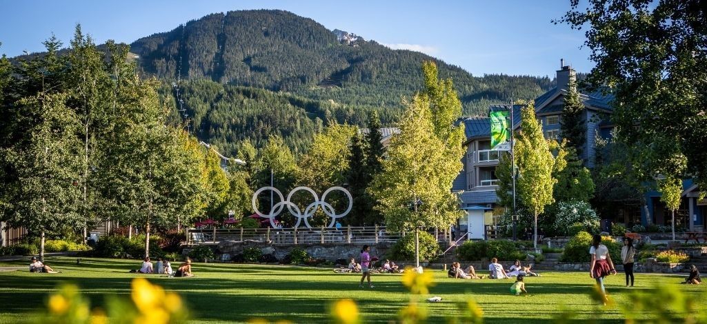 Whistler Olympic Plaza with green lawn, Olympic rings, surrounding trees and mountain views on a sunny summer day.