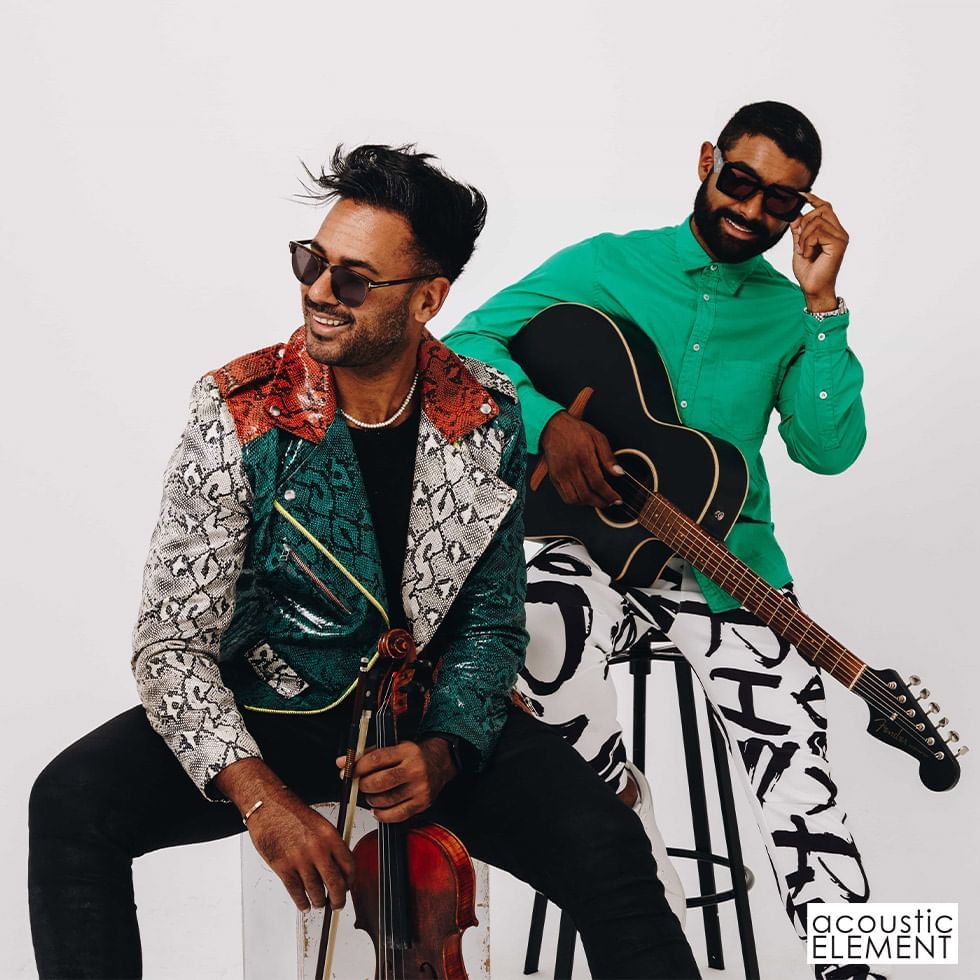 Two men sit together, one with a violin, the other with a guitar, both wearing sunglasses.