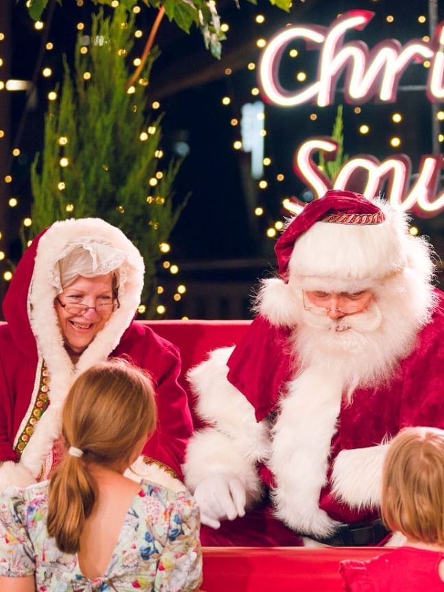 Children meeting Santa and Mrs. Claus on a festive red bench surrounded by twinkling lights and greenery.