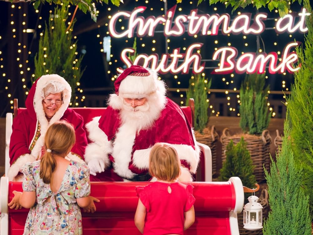 Children meeting Santa and Mrs. Claus on a festive red bench surrounded by twinkling lights and greenery.