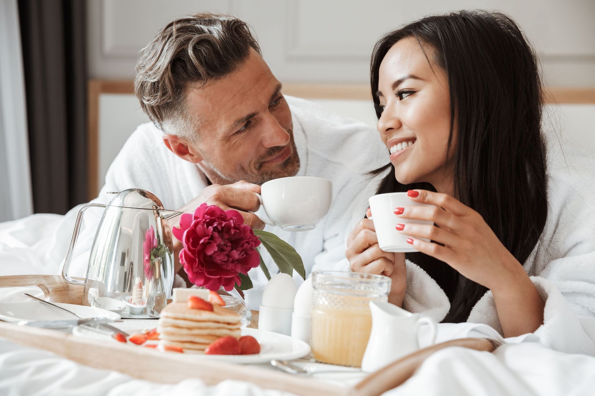 Couple in robes snuggling and smiling as they drink tea and eat 