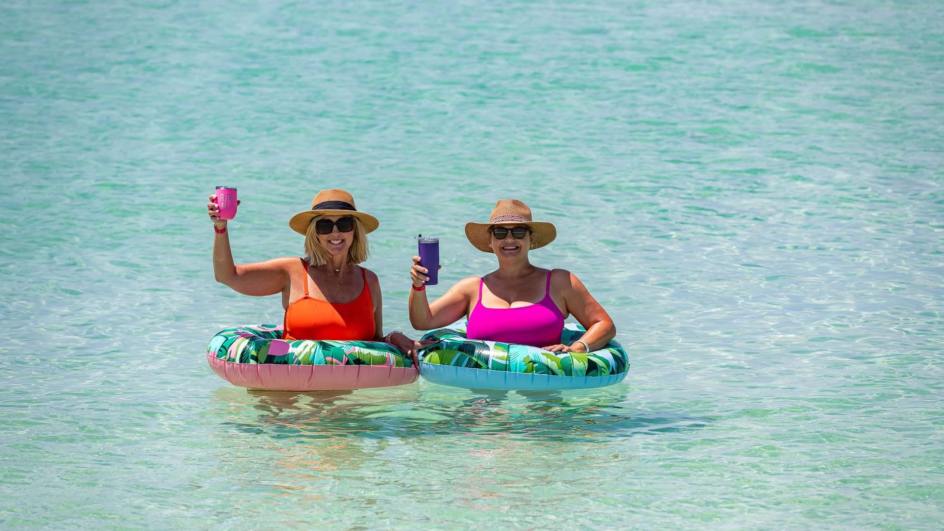 Two ladies raising a cup to cheers while wading in the gulf water with colorful inner tubes
