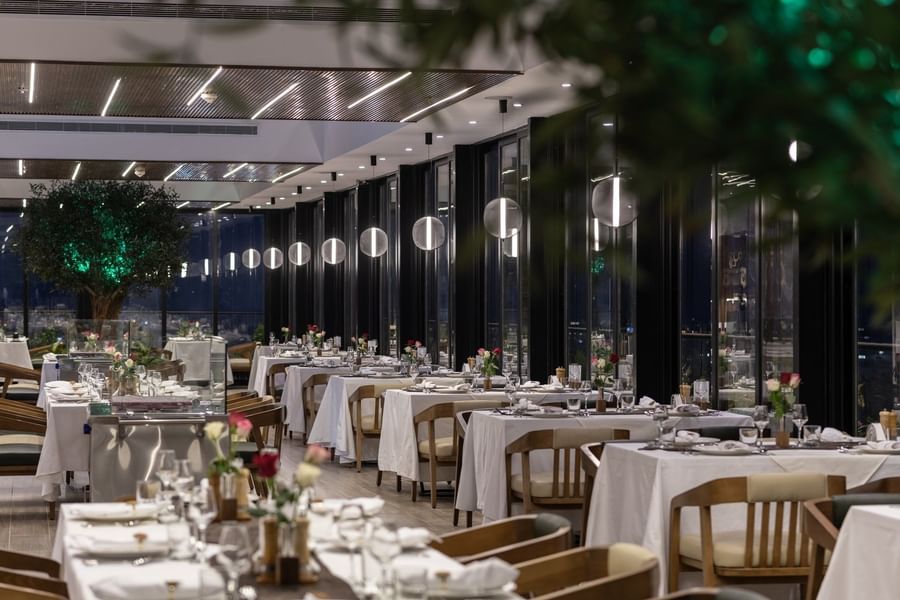 Karkan Restaurant dining area with white tables by glass walls under glowing globe lights at night at Warwick Al Taif Hotel