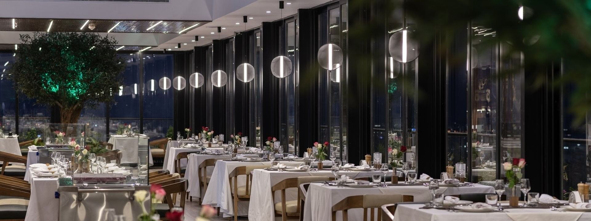 Karkan Restaurant dining area with white tables by glass walls under glowing globe lights at night at Warwick Al Taif Hotel
