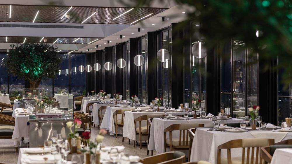 Karkan Restaurant dining area with white tables by glass walls under glowing globe lights at night at Warwick Al Taif Hotel