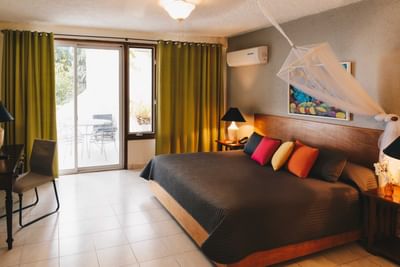 King bed with vibrant pillows under a canopy net, by green curtains & patio doors in Executive Suite at Hotel Montana Haiti
