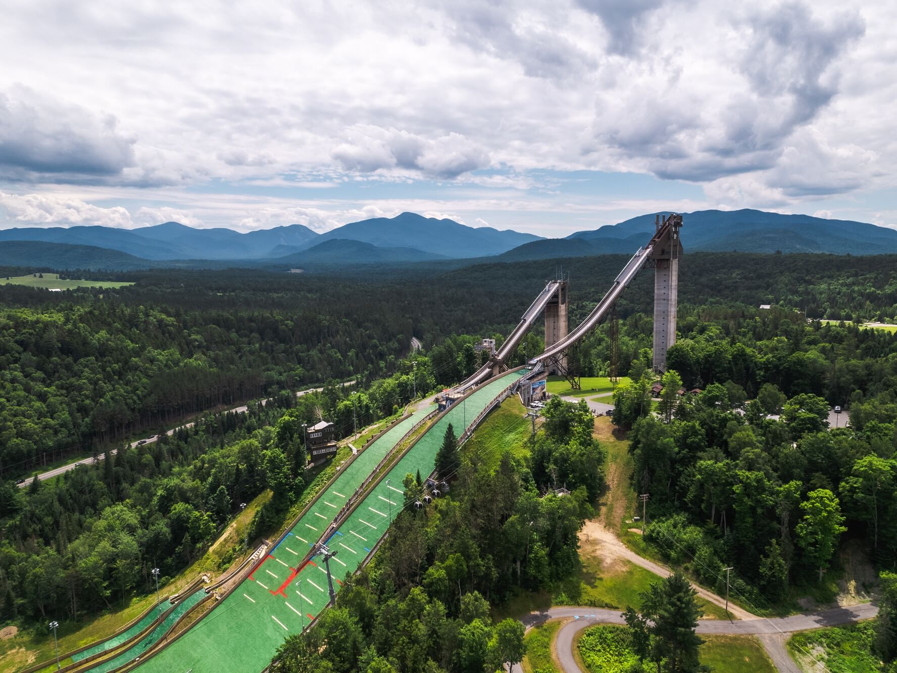 An aerial view shows the imposing Olympic ski jumping complex surrounded by green forests near Lake Placid Club Grandview