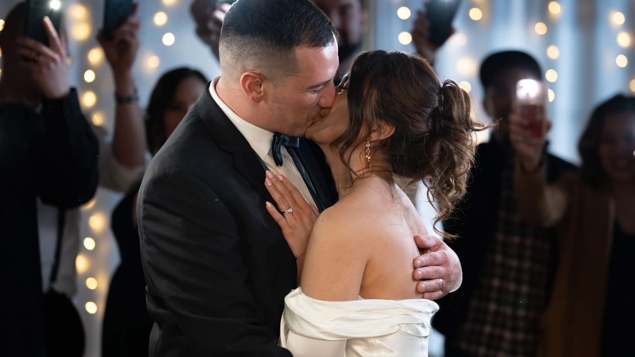 Man and woman in formal attire kiss as others watch and take photos with lights in background.