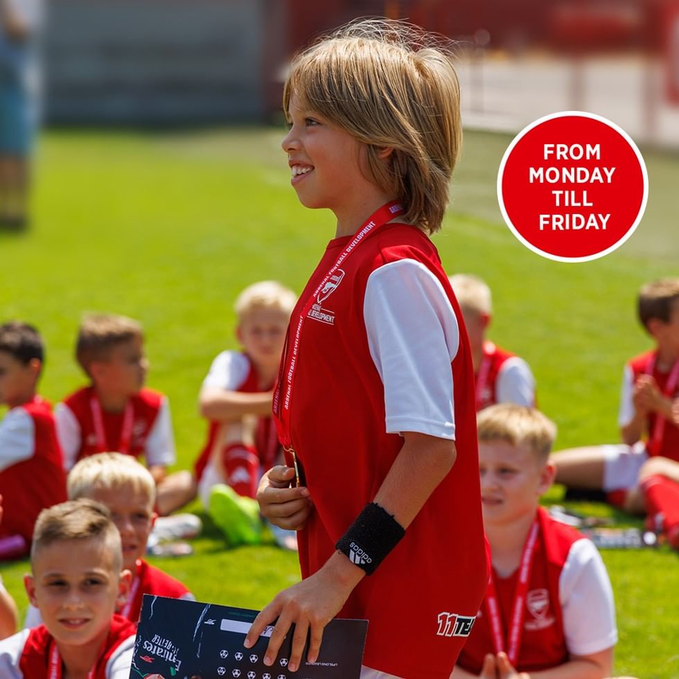 Lächelnder Junge in rot-weißer Fußballkleidung beim Arsenal Football Camp auf grünem Rasen mit anderen Kindern