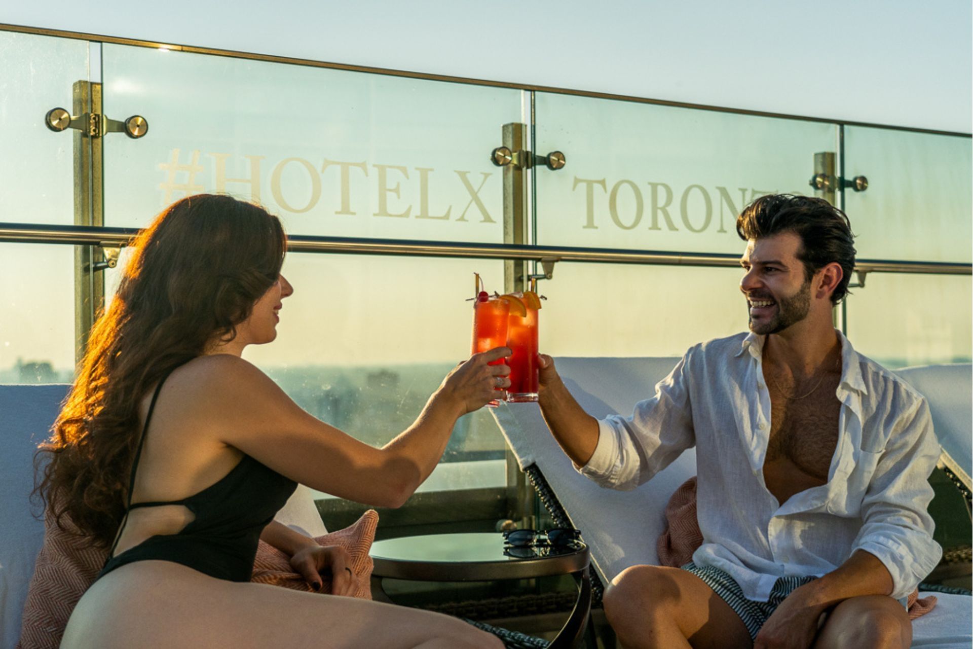 Couple toasting red cocktails on cushioned rooftop seats by a glass railing under warm sunset light at Hotel X Toronto