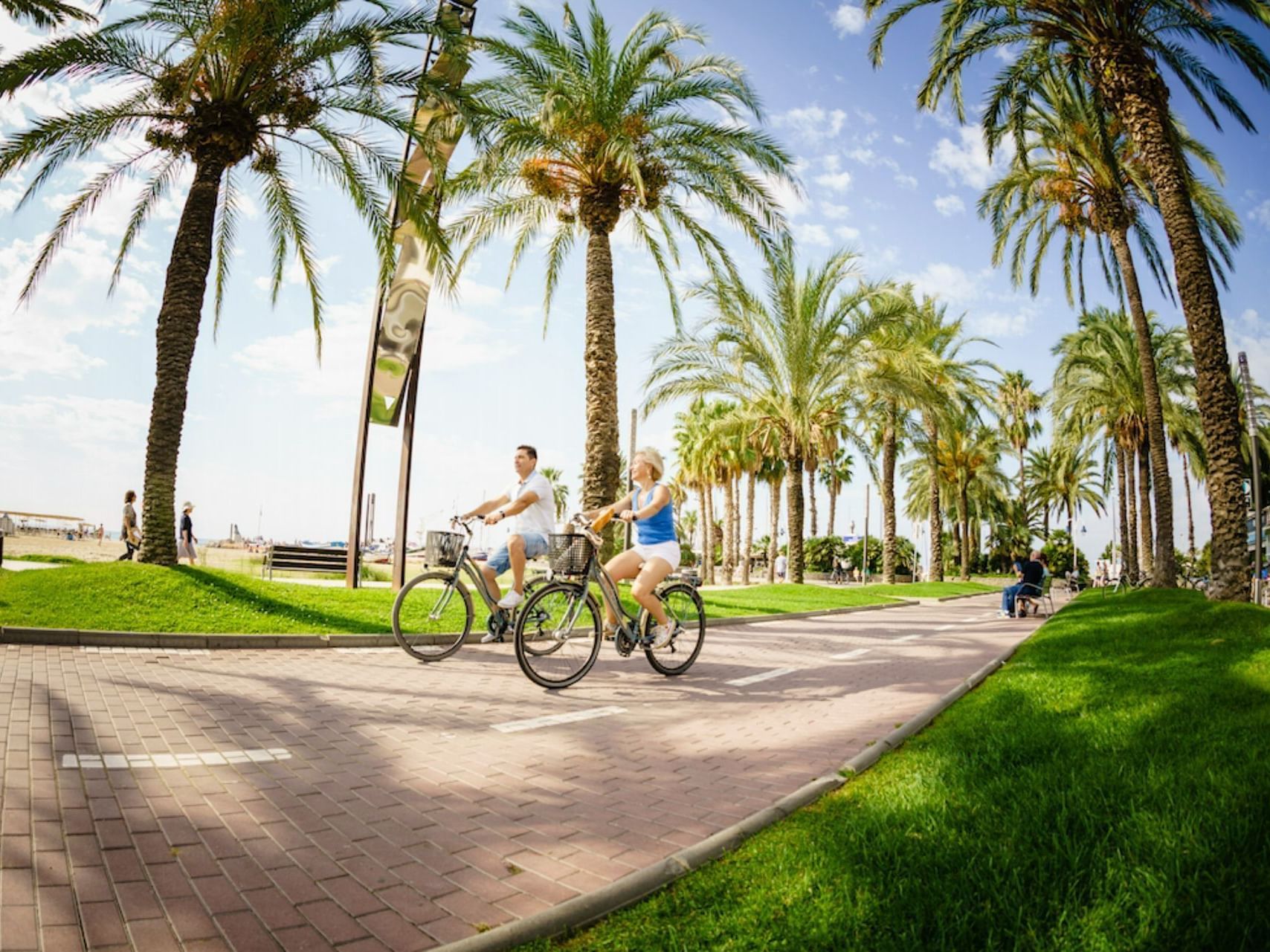 Two cyclists riding on a path surrounded by palm trees near Ponient Dorada Palace