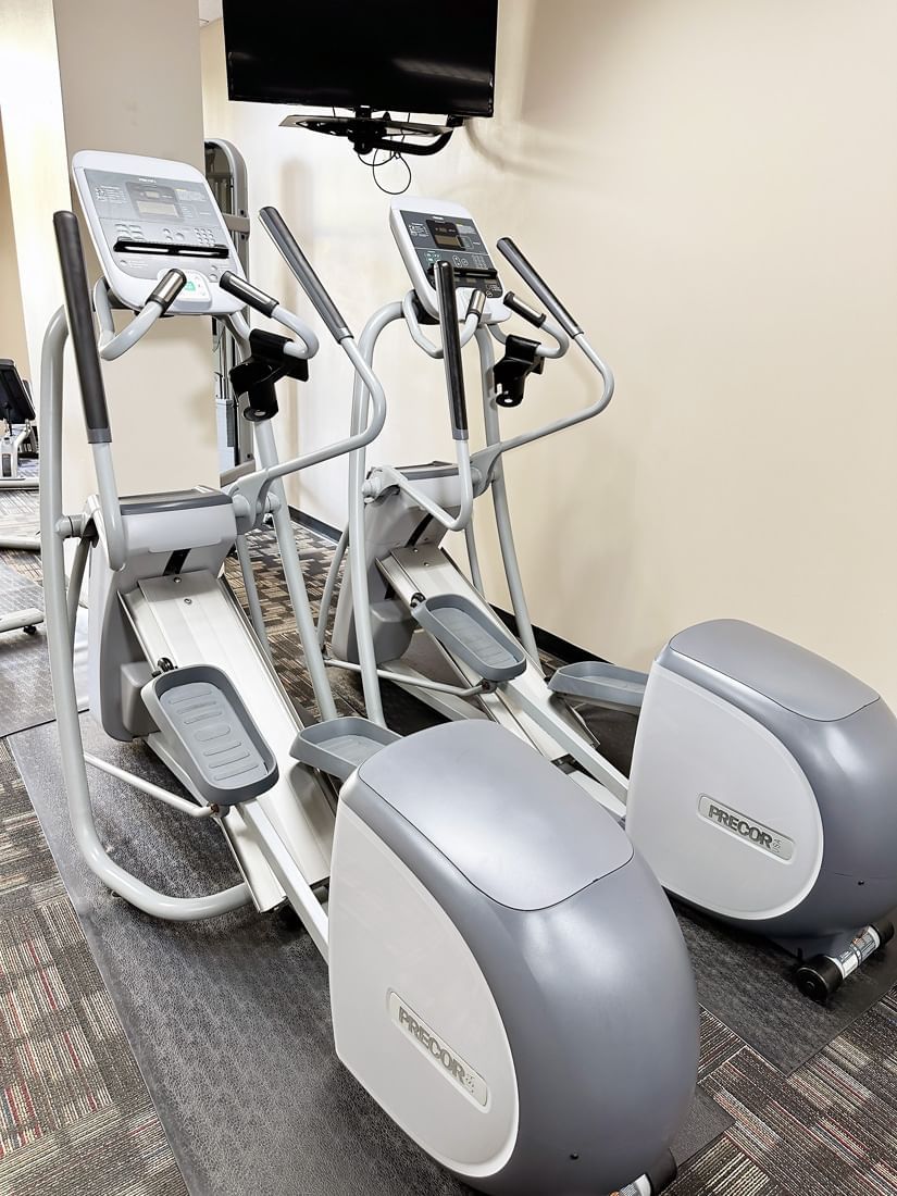 Two gray and white elliptical machines sit in a gym with a TV mounted on the wall.