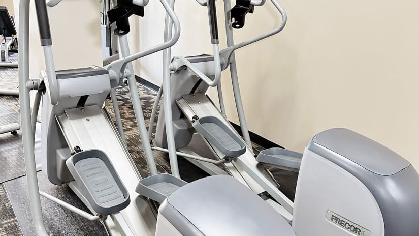 Two gray elliptical machines in a fitness center with gray carpeting and beige walls.