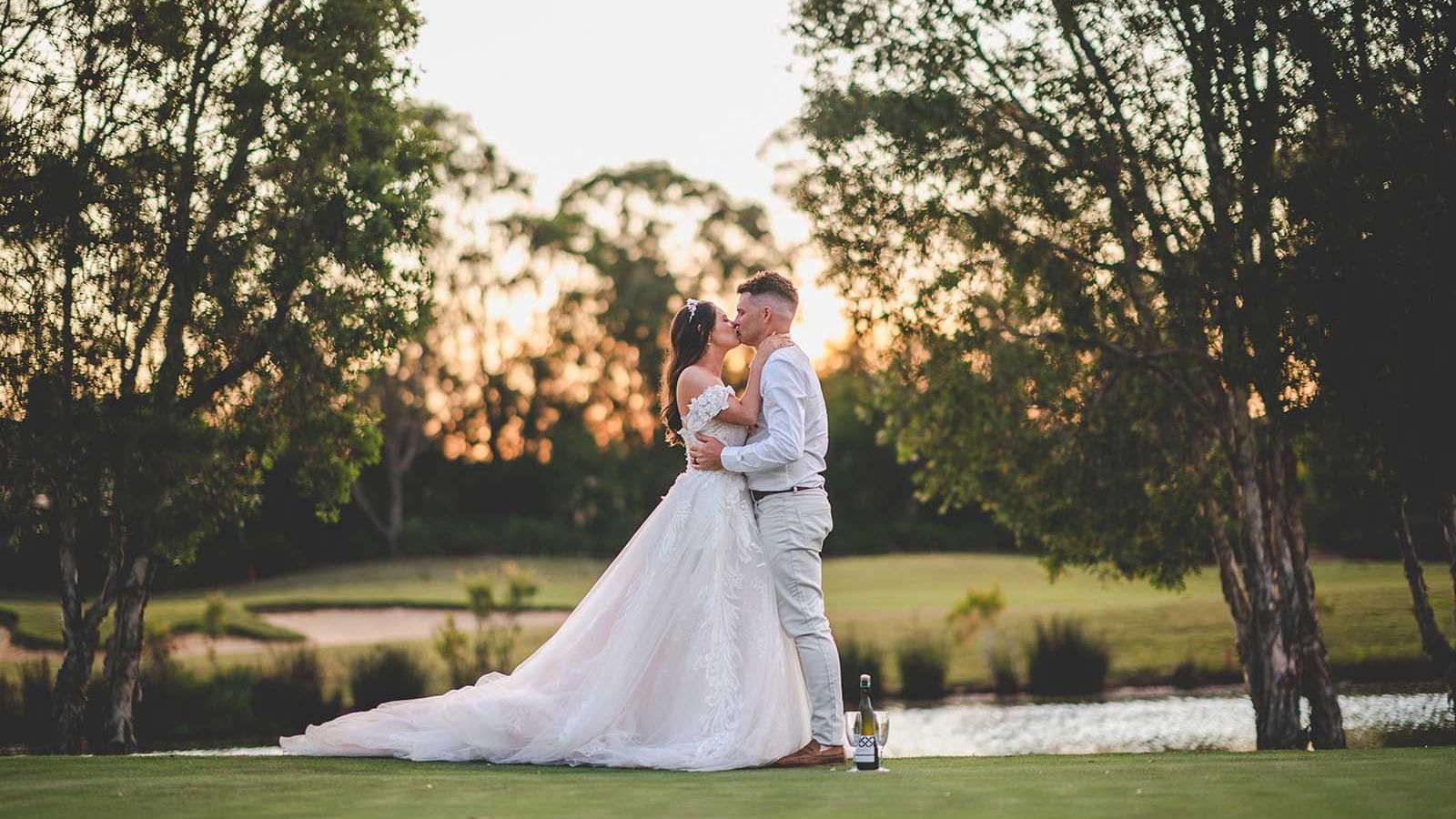 A bride and groom embrace by a lake at sunset, surrounded by trees near Mercure Kooindah Waters