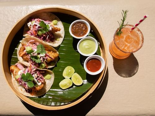 Fish tacos with sauces and a cocktail on a bamboo platter served at Sirenas Restaurant in Hacienda Del Mar Los Cabos.