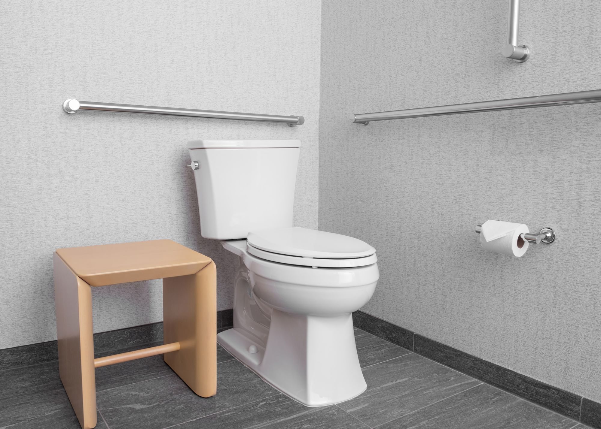 A standard bathroom with a toilet, stool, grab bars, and tile floor.
