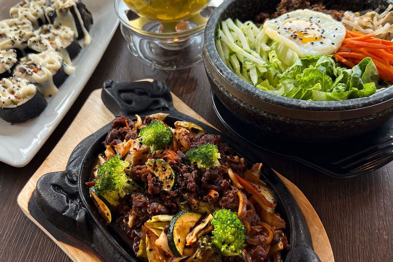 Bowl of bibimbap, pan of beef with vegetables, and plate of sushi rolls on a table.