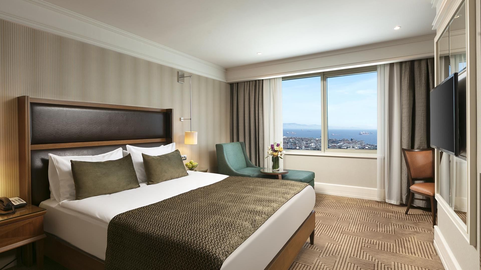 A hotel room with a large bed, two armchairs, a TV, and a large window overlooking the city in Deluxe Room at Titanic Port Bakırköy