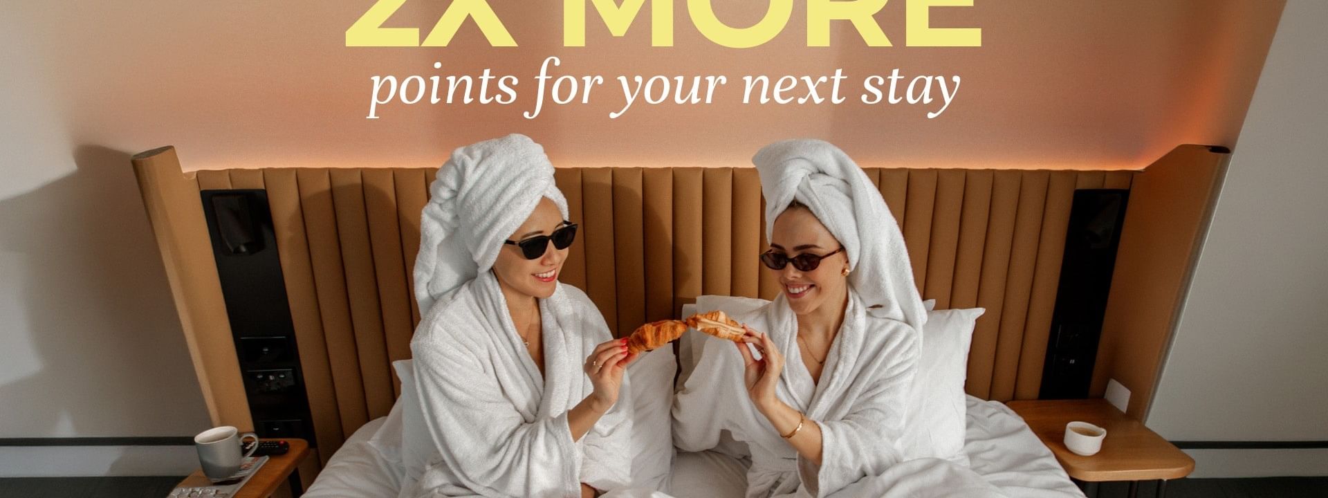 Two women in white robes and towels enjoying breakfast in bed with croissants for Double the Points on Your Next Stay.