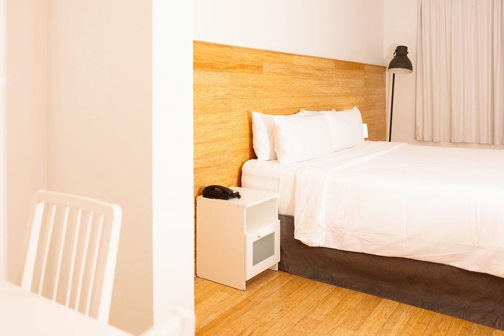 A hotel room with a king-sized bed, wood headboard, white linens, and a nightstand with a phone.
