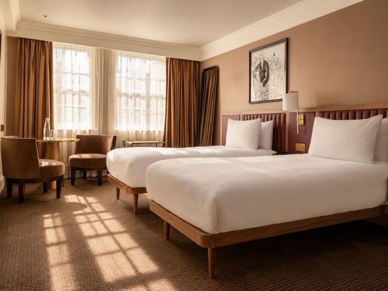 Bright Collection Deluxe Twin room with two beds, brown upholstered headboards, seating area, and large windows with drapes at The May Fair Hotel, London
