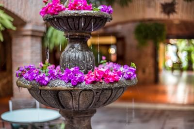 A stone fountain with water dripping and purple flowers, situated near a table and chairs.