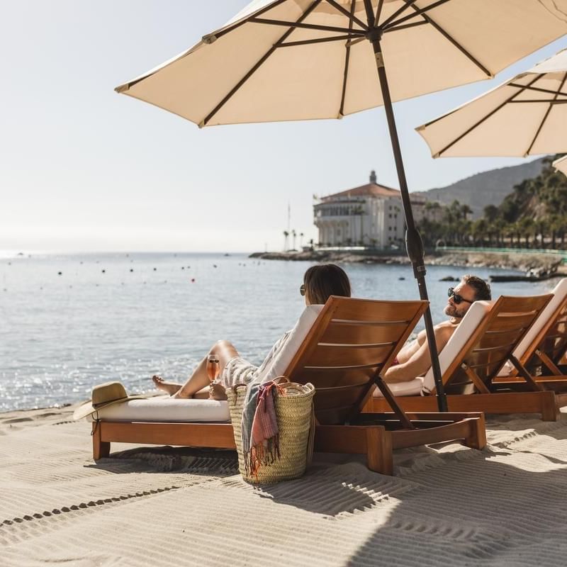 People relaxing on beach lounge chairs under umbrellas by the ocean.