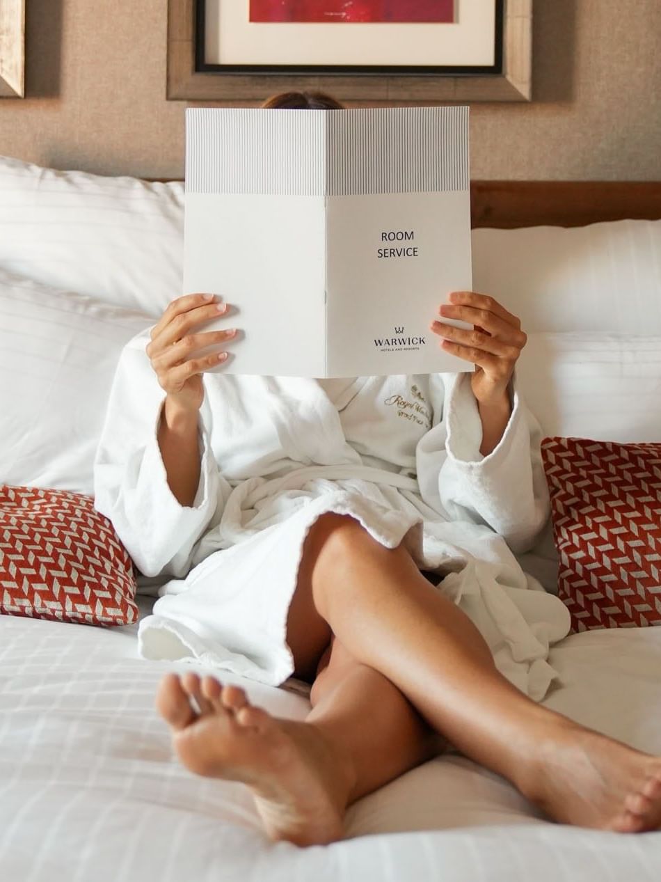 Guest in a bathrobe reading a room service menu placed by pillows on a bed at the Warwick Grand Place Brussels