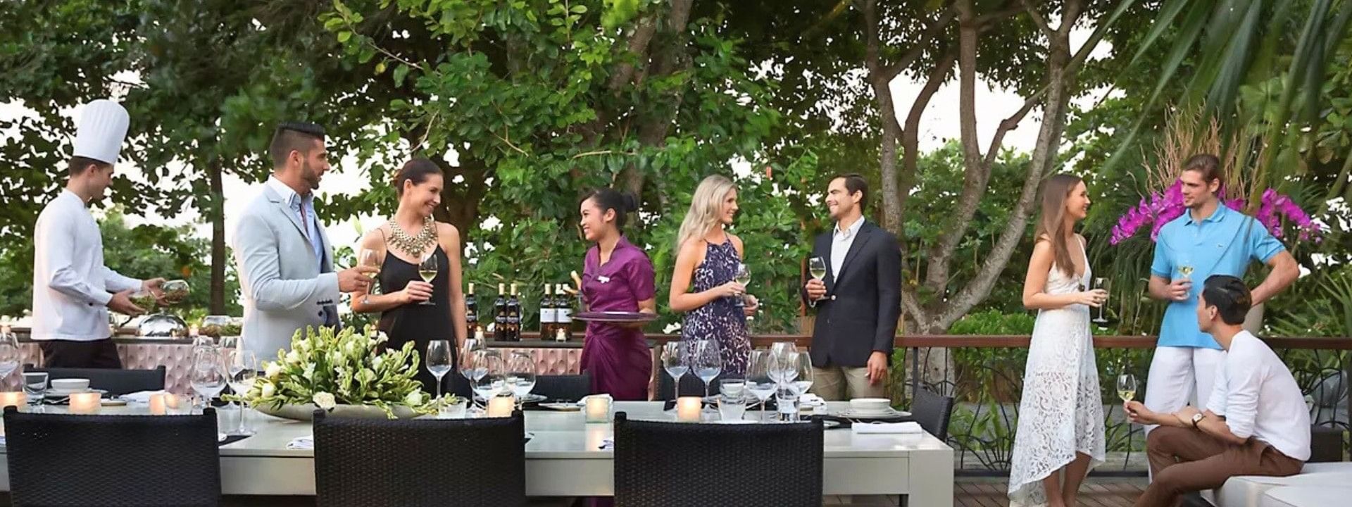 Group of people socializing on an outdoor deck with a dining table and lush greenery in the background.