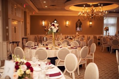 Elegantly set dining tables & chairs with tall floral centerpieces, a chandelier in the wedding hall at The Simsbury Inn