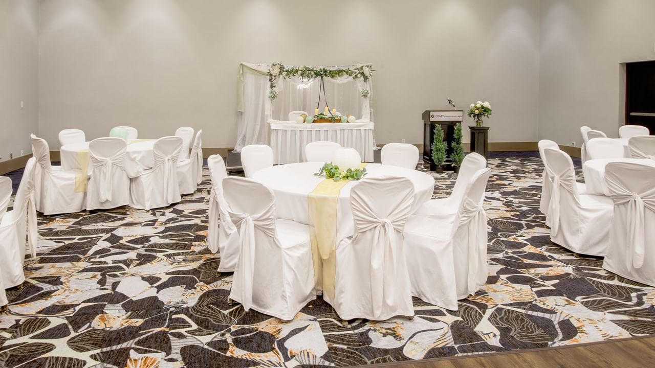 Banquet hall with round tables, draped chairs, and a decorated stage for a wedding.