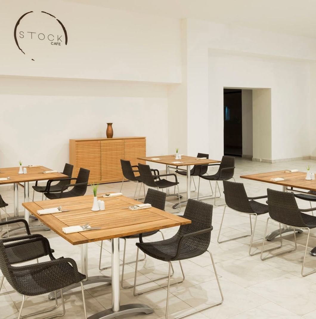 Black wicker chairs by wooden tables & hanging logo on a white wall in the Stock Cafe at Real Inn Mexicali