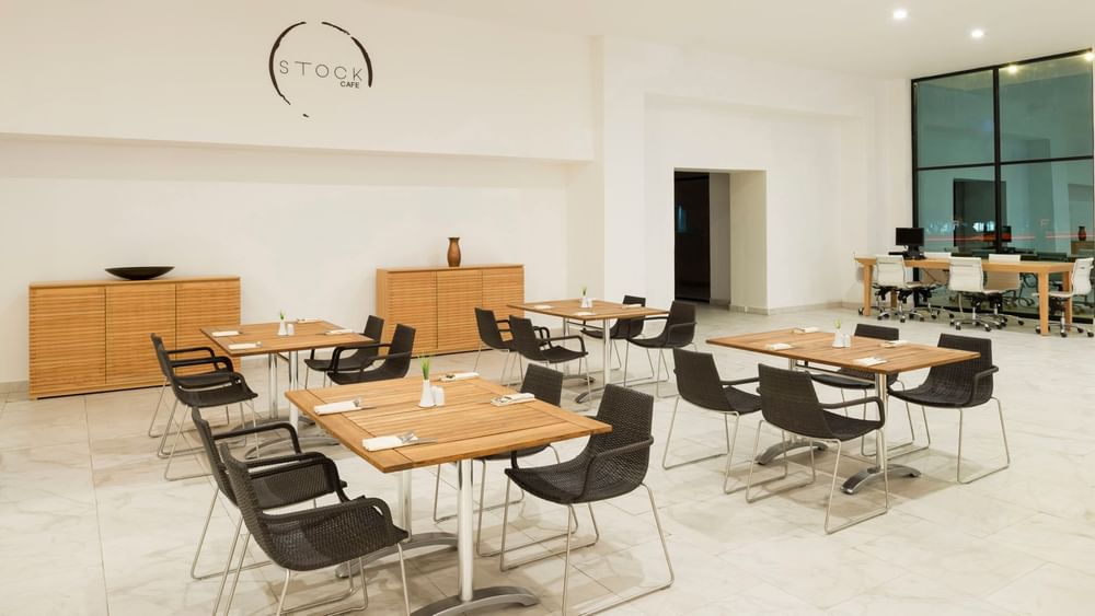 Black wicker chairs by wooden tables & hanging logo on a white wall in the Stock Cafe at Real Inn Mexicali