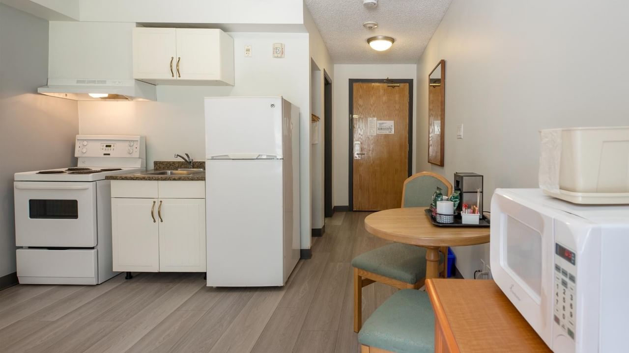 A kitchen with white appliances, wooden table, two chairs, and a microwave on a counter.