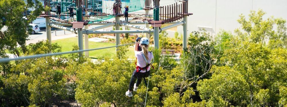 Woman zips across a zipline, wearing a helmet and harness, with an aerial obstacle course near Novotel Sunshine Coast Resort