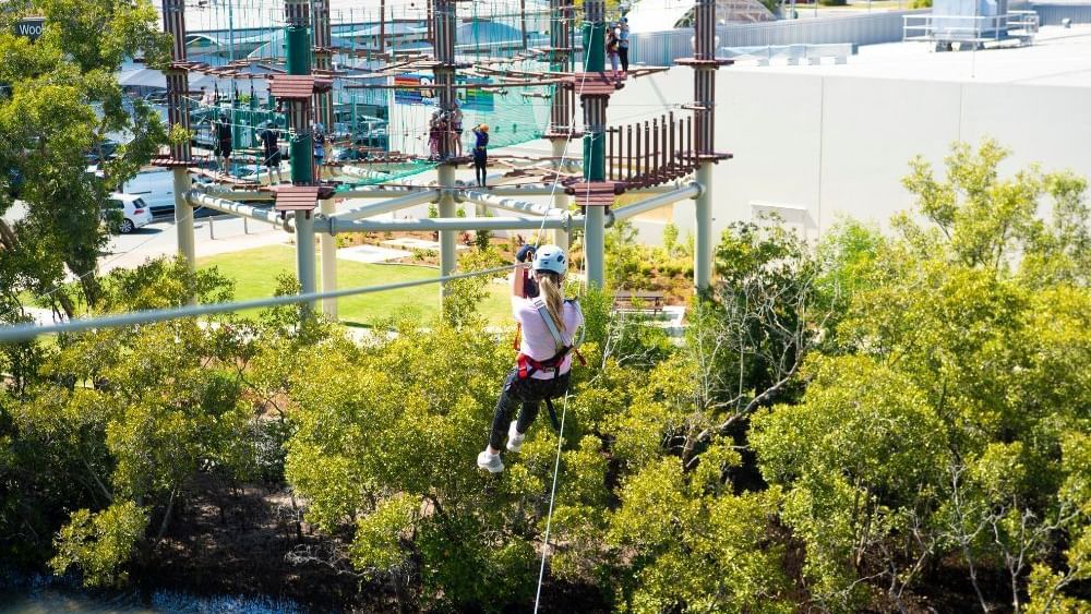 Woman zips across a zipline, wearing a helmet and harness, with an aerial obstacle course near Novotel Sunshine Coast Resort