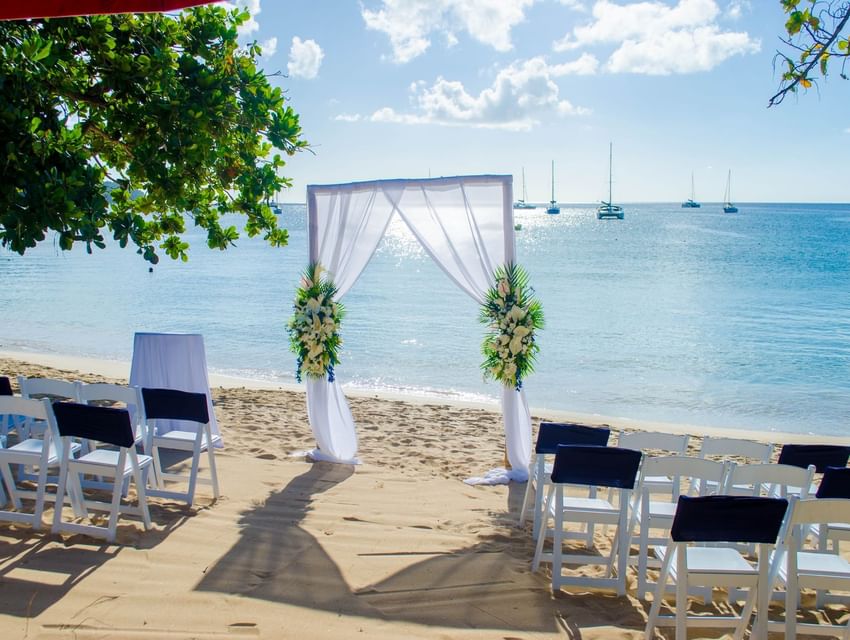Beach wedding setup with floral arch and white chairs adorned with green sashes at Bay Gardens Beach Resort and Spa