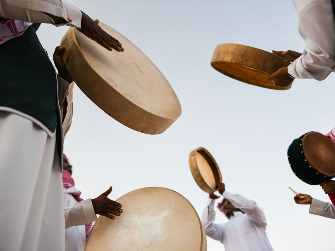 People in traditional Saudi clothing playing large hand drums outdoors at Saja Warwick Madinah