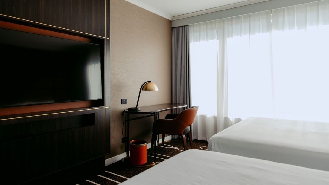 A hotel room with two beds, a desk with a lamp, and a large TV mounted on the wall.