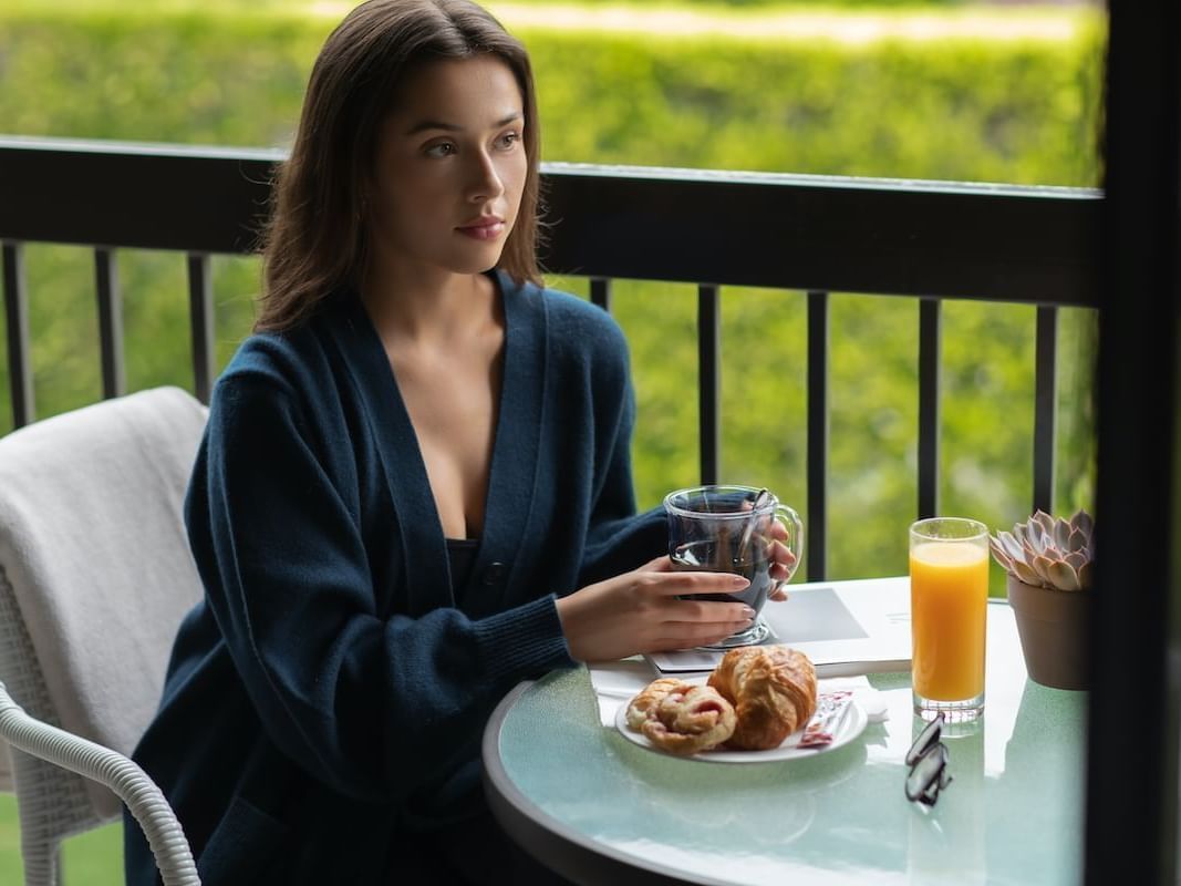 Woman wearing a cardigan enjoys breakfast on a private balcony at Luxe Sunset Boulevard Hotel