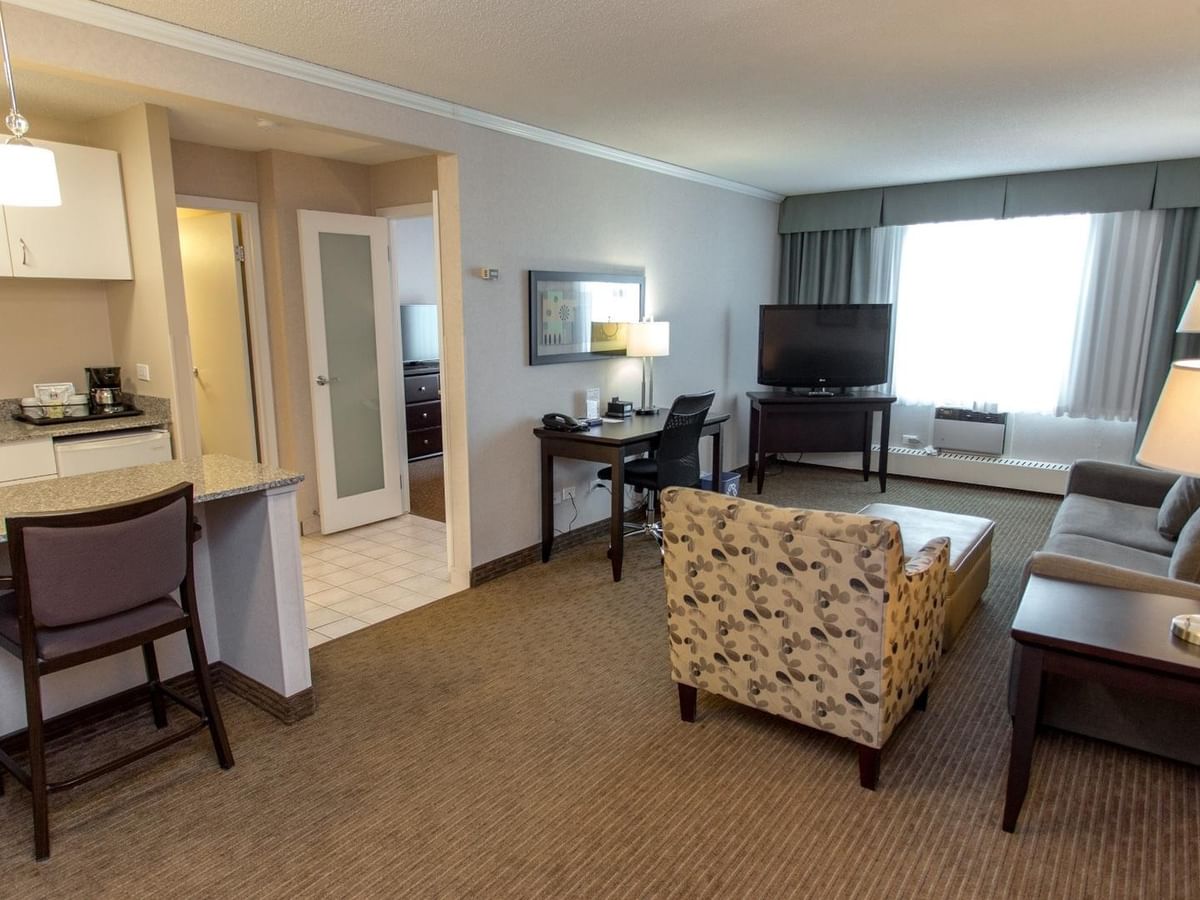 Hotel suite living area with a kitchenette, a bar, sofa, armchair, TV and work desk.
