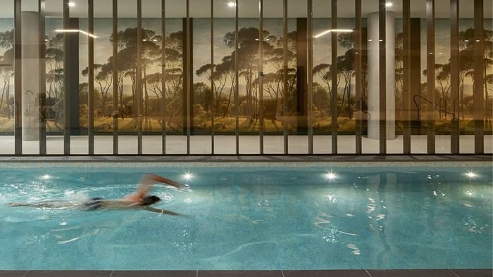 Man swimming in indoor pool with large, illuminated wall mural of a forest landscape.