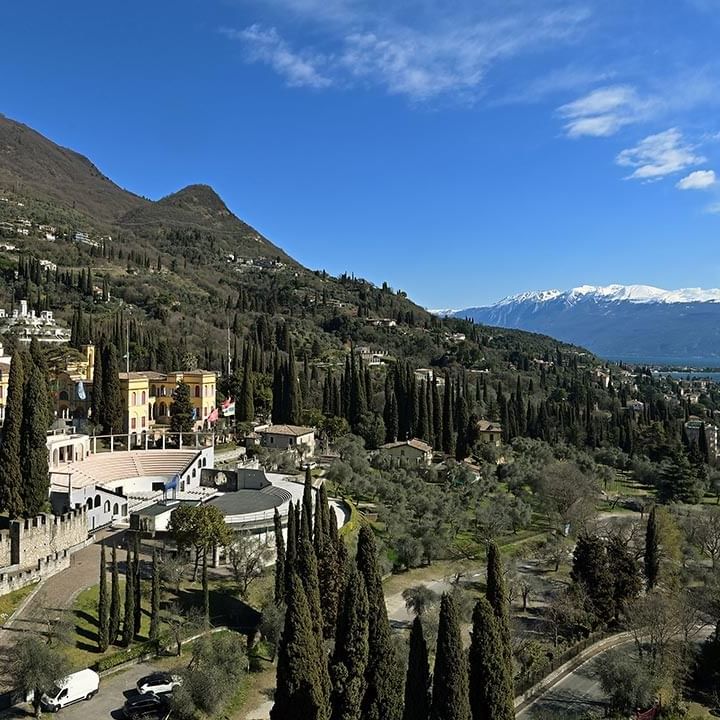 Vista panoramica di un'area collinare con edifici, cipressi, strada e acqua in lontananza.