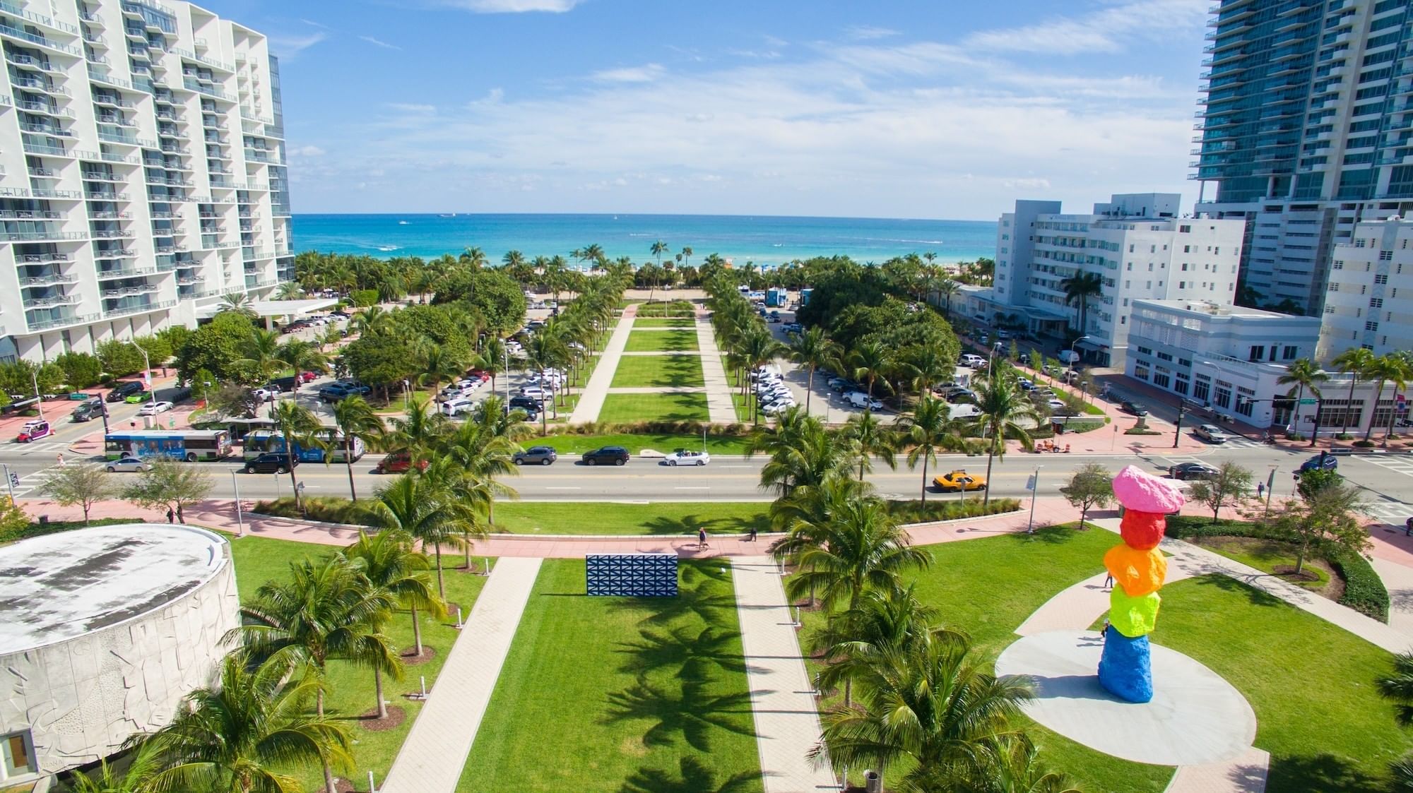 An aerial view of a park with palm trees, a sculpture, and a beach in the background.