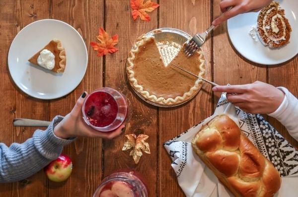 An overhead shot of a Thanksgiving table with pumpkin pie, autumnal leaves, braided bread, and someone toasting with a glass of wine.