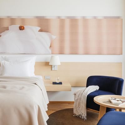 Camera da letto con letti fatti, comodini, lampade, poltrone blu, tavolino e grande quadro.